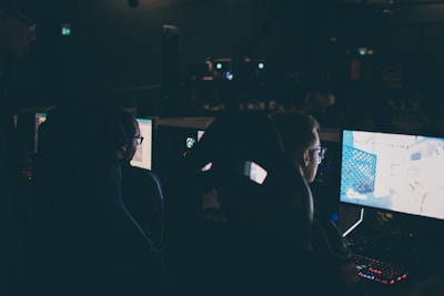 Several individuals are sitting in front of computer screens in a dimly lit room, likely engaging in gaming or working on tasks. The glow of the monitors illuminates their faces, creating a focused and immersive atmosphere. The surroundings suggest a setting like a gaming event or a LAN party.