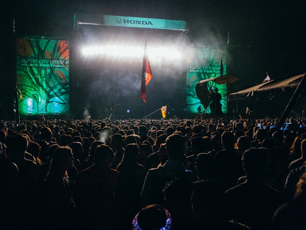 A large outdoor concert stage is illuminated with bright lights and flanked by vibrant banners that read 'Austin City Limits Music Festival.' The audience is densely packed, with people standing close together, many holding flags. The night atmosphere is evident, with dark surroundings except for the brightly lit stage.