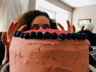 A large round cake covered in pink frosting with blueberries neatly lined along the top edge. Behind the cake, a person with wide eyes and curly hair peeks over, creating a playful and slightly mischievous atmosphere.