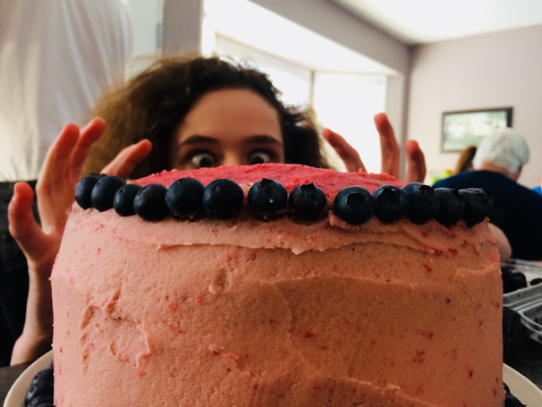 A large round cake covered in pink frosting with blueberries neatly lined along the top edge. Behind the cake, a person with wide eyes and curly hair peeks over, creating a playful and slightly mischievous atmosphere.
