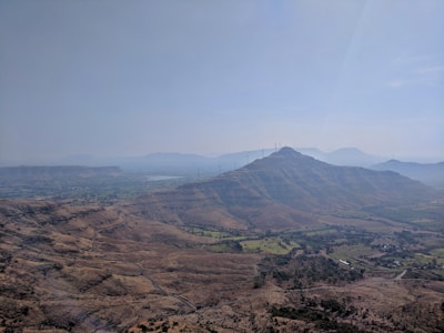 A scenic landscape showcasing a vast expanse of hilly terrain with scattered vegetation. A series of wind turbines is visible on the hilltops, emphasizing the presence of renewable energy infrastructure. The sky is clear, with a gentle haze in the distance, adding depth to the visual.