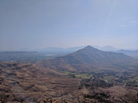 A scenic landscape showcasing a vast expanse of hilly terrain with scattered vegetation. A series of wind turbines is visible on the hilltops, emphasizing the presence of renewable energy infrastructure. The sky is clear, with a gentle haze in the distance, adding depth to the visual.