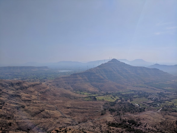A scenic landscape showcasing a vast expanse of hilly terrain with scattered vegetation. A series of wind turbines is visible on the hilltops, emphasizing the presence of renewable energy infrastructure. The sky is clear, with a gentle haze in the distance, adding depth to the visual.