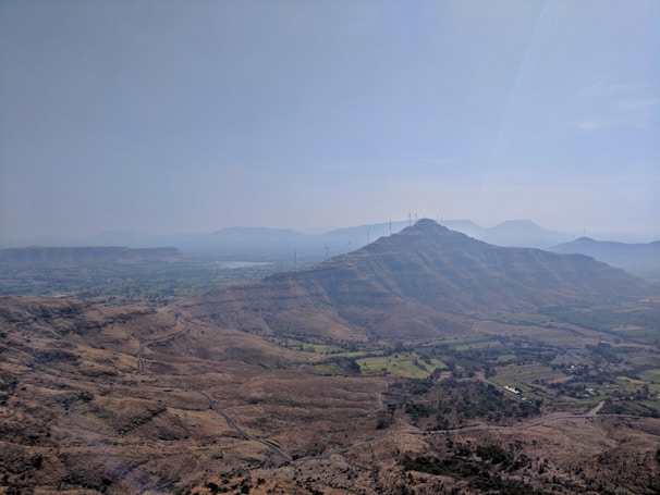 A scenic landscape showcasing a vast expanse of hilly terrain with scattered vegetation. A series of wind turbines is visible on the hilltops, emphasizing the presence of renewable energy infrastructure. The sky is clear, with a gentle haze in the distance, adding depth to the visual.