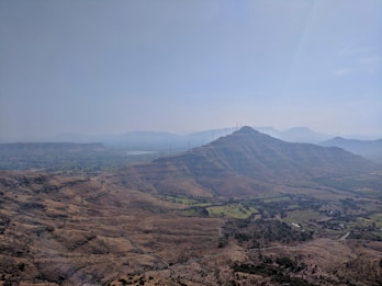 A scenic landscape showcasing a vast expanse of hilly terrain with scattered vegetation. A series of wind turbines is visible on the hilltops, emphasizing the presence of renewable energy infrastructure. The sky is clear, with a gentle haze in the distance, adding depth to the visual.