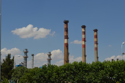 Several tall smokestacks with red and white bands rise above a lush green canopy of trees against a backdrop of a blue sky filled with fluffy white clouds.
