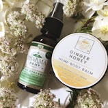 A flat lay composition showcases a dark glass bottle labeled 'Pharma Hemp' and a round container labeled 'Ginger Honey Hemp Body Balm,' surrounded by white flowers with green stems. The bottle appears to be a form of hemp oil, while the circular container is likely a skincare product.