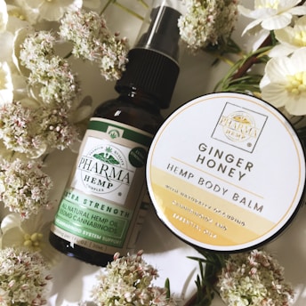 A flat lay composition showcases a dark glass bottle labeled 'Pharma Hemp' and a round container labeled 'Ginger Honey Hemp Body Balm,' surrounded by white flowers with green stems. The bottle appears to be a form of hemp oil, while the circular container is likely a skincare product.