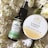A flat lay composition showcases a dark glass bottle labeled 'Pharma Hemp' and a round container labeled 'Ginger Honey Hemp Body Balm,' surrounded by white flowers with green stems. The bottle appears to be a form of hemp oil, while the circular container is likely a skincare product.