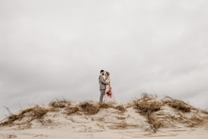 A couple dressed in wedding attire stands intimately close on a sand dune surrounded by sparse vegetation. The sky is overcast with a muted color palette, adding a serene and romantic ambiance.