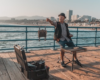 A street performer is engaging with an audience on a pier with the ocean in the background. The performer, wearing a hat and a vest, is pointing outward, while small props are visible on the table in front of him. A suitcase filled with equipment is open on the ground. In the distance, a city skyline and mountains are visible.