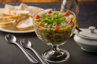 A glass dish filled with a colorful salad featuring chopped herbs, red pepper pieces, and garnished with mint leaves. It is set on a black textured mat alongside two spoons. In the background, there is a plate with sliced bread and a small white dish with a lid.