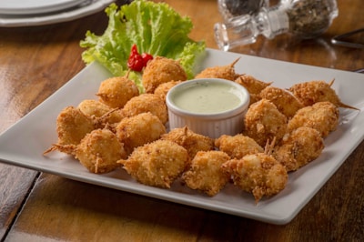 A rectangular white plate is filled with crispy, golden-brown fried appetizers arranged in a circular pattern around a small cup of creamy dipping sauce. The dish is garnished with fresh lettuce leaves and a single red decorative vegetable. In the background, there are salt and pepper shakers on a wooden table surface.
