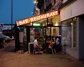 An outdoor restaurant with a brightly lit sign reading 'BAR RESTAURANT' under a dusky sky. Several people are sitting at tables inside a glass-enclosed area, engaged in conversation. There's a menu board leaning against the wall, and the reflections in the glass create a cozy ambiance.