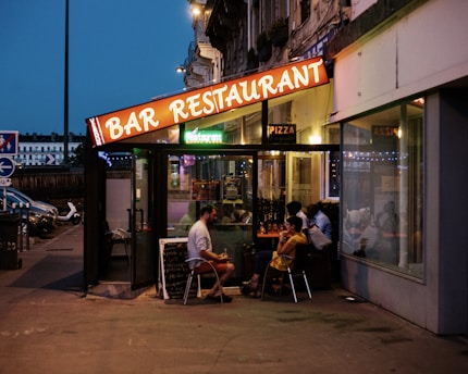 An outdoor restaurant with a brightly lit sign reading 'BAR RESTAURANT' under a dusky sky. Several people are sitting at tables inside a glass-enclosed area, engaged in conversation. There's a menu board leaning against the wall, and the reflections in the glass create a cozy ambiance.