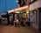 An outdoor restaurant with a brightly lit sign reading 'BAR RESTAURANT' under a dusky sky. Several people are sitting at tables inside a glass-enclosed area, engaged in conversation. There's a menu board leaning against the wall, and the reflections in the glass create a cozy ambiance.