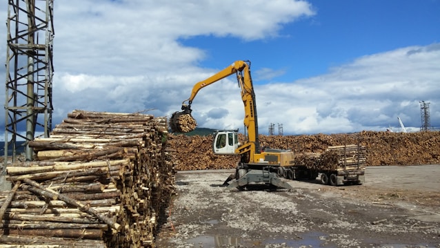 A large industrial crane with a yellow arm is stacking logs at a lumber yard. Piles of cut logs are arranged on the ground, and a truck loaded with timber is visible. The sky is partly cloudy, providing a bright backdrop to the scene.