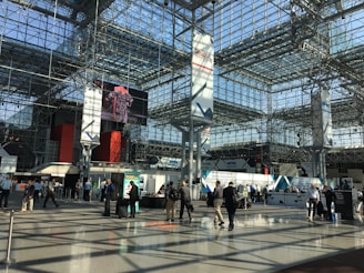 A spacious, modern convention center with a high, glass ceiling. The interior structure is made up of metal beams and large pillars. Several people are walking or gathering in small groups, some standing near exhibition booths and registration counters. A large digital screen displays an image above the main floor.