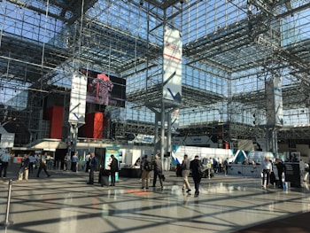 A spacious, modern convention center with a high, glass ceiling. The interior structure is made up of metal beams and large pillars. Several people are walking or gathering in small groups, some standing near exhibition booths and registration counters. A large digital screen displays an image above the main floor.