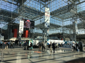A spacious, modern convention center with a high, glass ceiling. The interior structure is made up of metal beams and large pillars. Several people are walking or gathering in small groups, some standing near exhibition booths and registration counters. A large digital screen displays an image above the main floor.