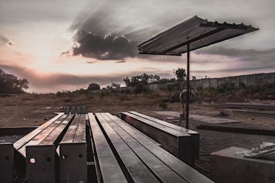 Steel beams are neatly arranged on the ground in an open, desolate area with a metal canopy structure providing partial cover. The setting is outdoor, with a dramatic sky capturing a cloud partially covering the sun, creating sunbeams that spread across the sky. In the background, there are trees and a tall concrete wall.
