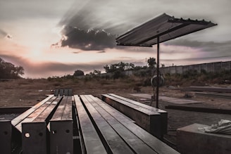 Steel beams are neatly arranged on the ground in an open, desolate area with a metal canopy structure providing partial cover. The setting is outdoor, with a dramatic sky capturing a cloud partially covering the sun, creating sunbeams that spread across the sky. In the background, there are trees and a tall concrete wall.
