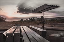 Steel beams are neatly arranged on the ground in an open, desolate area with a metal canopy structure providing partial cover. The setting is outdoor, with a dramatic sky capturing a cloud partially covering the sun, creating sunbeams that spread across the sky. In the background, there are trees and a tall concrete wall.