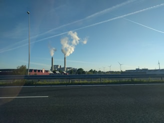 Two industrial chimneys release white smoke into the clear blue sky. A wide road with a speed limit sign is in the foreground, accompanied by several wind turbines spread across the landscape. The horizon features a combination of buildings, vegetation, and a few clouds overhead.