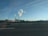 Two industrial chimneys release white smoke into the clear blue sky. A wide road with a speed limit sign is in the foreground, accompanied by several wind turbines spread across the landscape. The horizon features a combination of buildings, vegetation, and a few clouds overhead.
