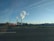 Two industrial chimneys release white smoke into the clear blue sky. A wide road with a speed limit sign is in the foreground, accompanied by several wind turbines spread across the landscape. The horizon features a combination of buildings, vegetation, and a few clouds overhead.