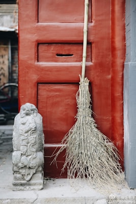 A rustic broom made from straw is leaning against a large, red wooden door. Next to the broom is an intricately carved stone statue, likely representing a mythical creature or guardian. The background suggests an old, possibly traditional setting.