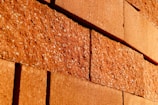 Rough textured red bricks with visible specks are stacked in an offset pattern. The bricks are closely aligned and the lighting casts warm tones across the surface, highlighting the rugged surface details.