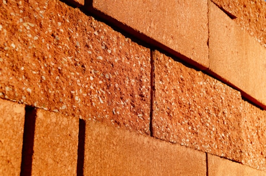 Rough textured red bricks with visible specks are stacked in an offset pattern. The bricks are closely aligned and the lighting casts warm tones across the surface, highlighting the rugged surface details.