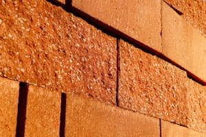 Rough textured red bricks with visible specks are stacked in an offset pattern. The bricks are closely aligned and the lighting casts warm tones across the surface, highlighting the rugged surface details.