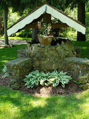 A rustic stone well adorned with a wooden roof structure stands in a lush garden. Hanging from the center is a basket overflowing with vibrant pink flowers. Around the base, hostas with variegated green and white leaves are planted, surrounded by manicured grass and shaded by tall trees.