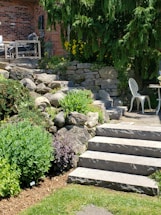A landscaped garden area featuring stone steps leading up to a brick facade of a house. There are lush green shrubs and plants lining the stone path, with yellow flowers adding a pop of color. A wooden outdoor seating set is partially visible, as well as a white metal chair and small circular table on the patio. Tall evergreens provide shade and backdrop to the scene.