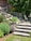 A landscaped garden area featuring stone steps leading up to a brick facade of a house. There are lush green shrubs and plants lining the stone path, with yellow flowers adding a pop of color. A wooden outdoor seating set is partially visible, as well as a white metal chair and small circular table on the patio. Tall evergreens provide shade and backdrop to the scene.