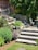 A landscaped garden area featuring stone steps leading up to a brick facade of a house. There are lush green shrubs and plants lining the stone path, with yellow flowers adding a pop of color. A wooden outdoor seating set is partially visible, as well as a white metal chair and small circular table on the patio. Tall evergreens provide shade and backdrop to the scene.