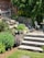 A landscaped garden area featuring stone steps leading up to a brick facade of a house. There are lush green shrubs and plants lining the stone path, with yellow flowers adding a pop of color. A wooden outdoor seating set is partially visible, as well as a white metal chair and small circular table on the patio. Tall evergreens provide shade and backdrop to the scene.