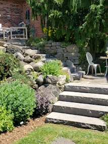 A landscaped garden area featuring stone steps leading up to a brick facade of a house. There are lush green shrubs and plants lining the stone path, with yellow flowers adding a pop of color. A wooden outdoor seating set is partially visible, as well as a white metal chair and small circular table on the patio. Tall evergreens provide shade and backdrop to the scene.
