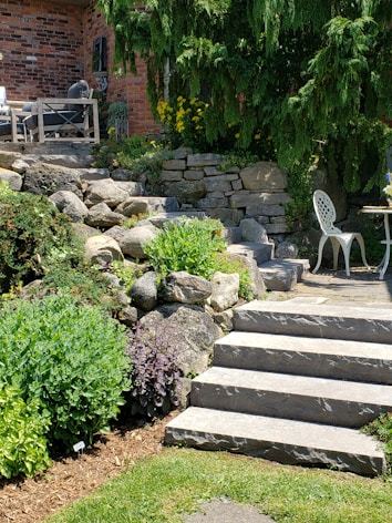 A landscaped garden area featuring stone steps leading up to a brick facade of a house. There are lush green shrubs and plants lining the stone path, with yellow flowers adding a pop of color. A wooden outdoor seating set is partially visible, as well as a white metal chair and small circular table on the patio. Tall evergreens provide shade and backdrop to the scene.