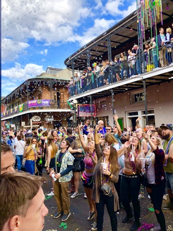 A lively street scene with a large crowd of people wearing colorful beads and festive attire, typical of a celebration or parade. People are reaching upwards, possibly catching or throwing beads. The scene features a two-story building with a balcony full of onlookers, decorated with banners. The sky is partly cloudy, suggesting daytime.