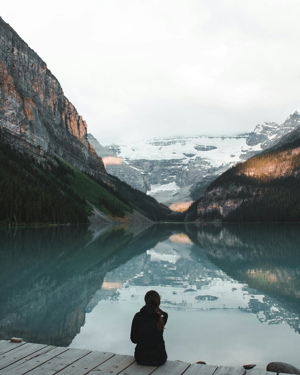 person sitting in a wooden floor near body of water and mountain during daytime