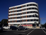 A modern multi-story building with a distinctive curved design features a pattern of horizontal white and dark panels with orange accents. Parked cars line the street in front, and several traffic lights are visible.