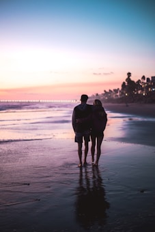 A couple walks along a tranquil beach at sunset, their figures silhouetted against the colorful horizon. The sky transitions from deep blue to soft pinks and purples, casting a gentle glow over the water. The beach is calm and reflects the vibrant colors of the sky.
