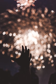 A silhouetted hand is raised against a backdrop of blurred golden fireworks. The image captures a sense of wonder and celebration.