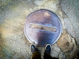 A manhole cover labeled 'City of Albany' is seen from above, embedded in a textured concrete surface. Two feet in sneakers are visible at the bottom of the frame, suggesting someone standing over the cover.