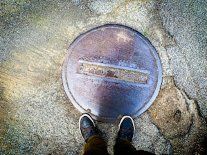 A manhole cover labeled 'City of Albany' is seen from above, embedded in a textured concrete surface. Two feet in sneakers are visible at the bottom of the frame, suggesting someone standing over the cover.