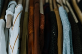 A row of various clothing items hanging on wooden hangers, featuring different textures and colors. The clothes include patterns and solid colors, predominantly in muted and warm tones.