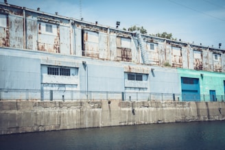 A weathered industrial building with rusty metal siding alongside a concrete waterfront. There are multiple windows and an exterior staircase. The building's exterior shows signs of decay with rust marks and peeling paint. The water in the foreground is calm, bordered by a concrete embankment.